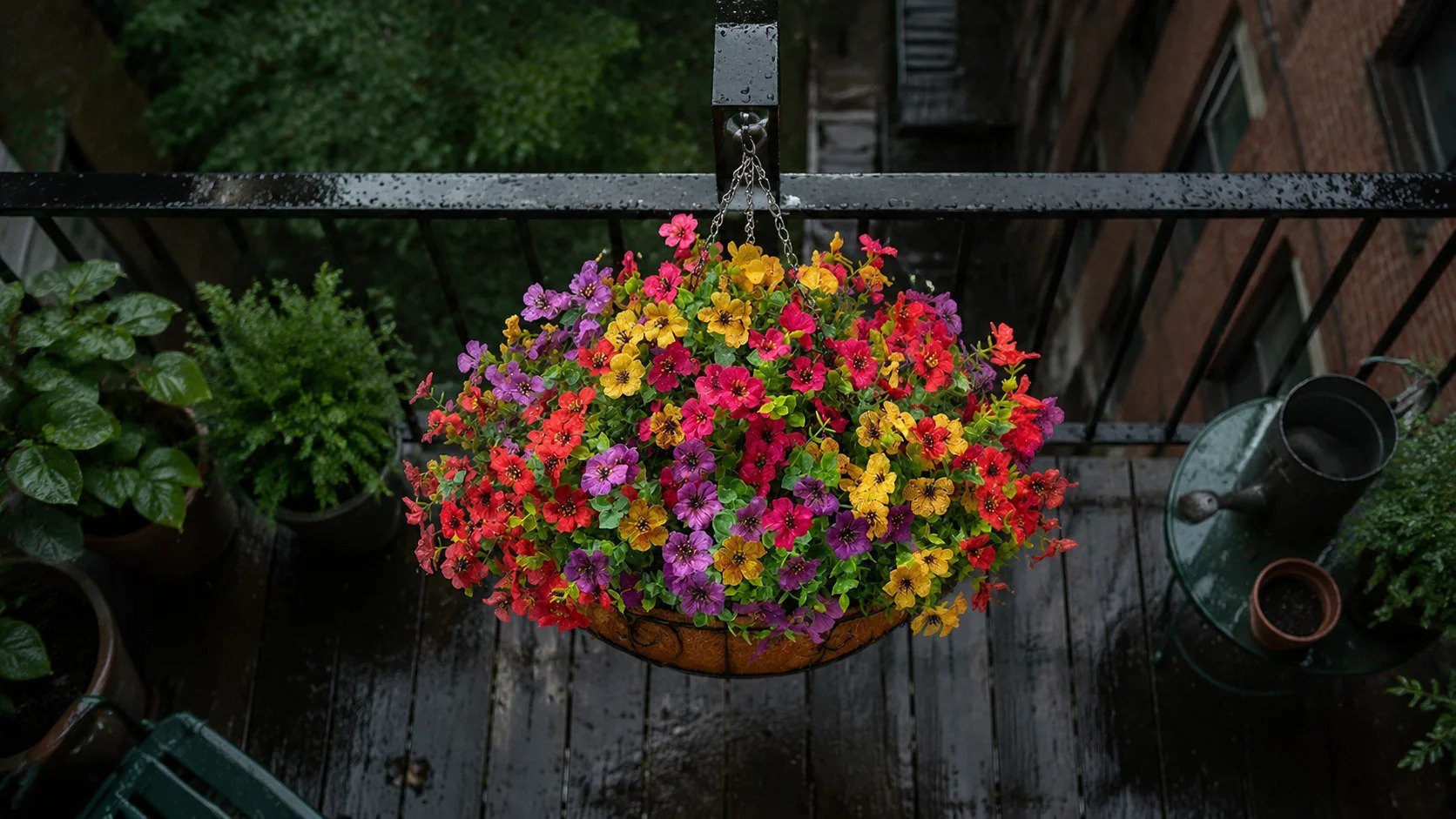 Overhead shot of a smooth cone-shaped terracotta hanging basket with purple, yellow, and deep orange UV resistant fake daisies and faux eucalyptus, glistening with raindrops on a wet Brooklyn balcony during a soft rain shower