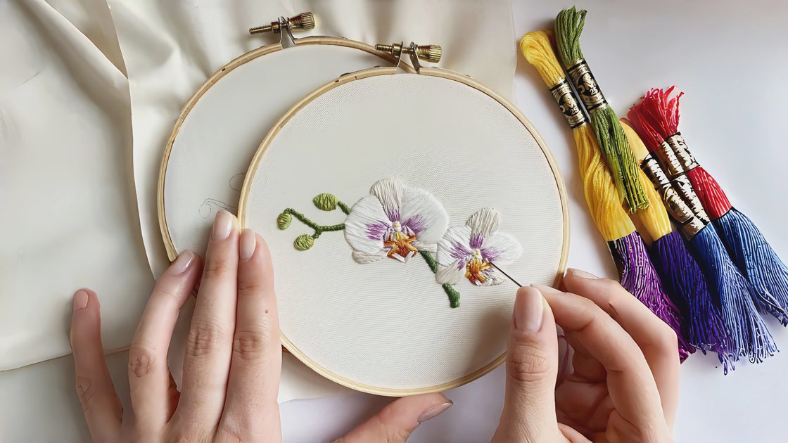 Hand embroidering a small pink floral cluster on a shirt collar using a large eye sewing needle, with colorful embroidery floss and a transparent needle storage tube nearby.
