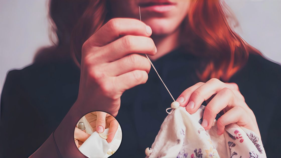 A hand pulling a large eye sewing needle with cream thread through a loose button on a wool coat, with a bright yellow needle storage tube and threader resting on a wooden table nearby.